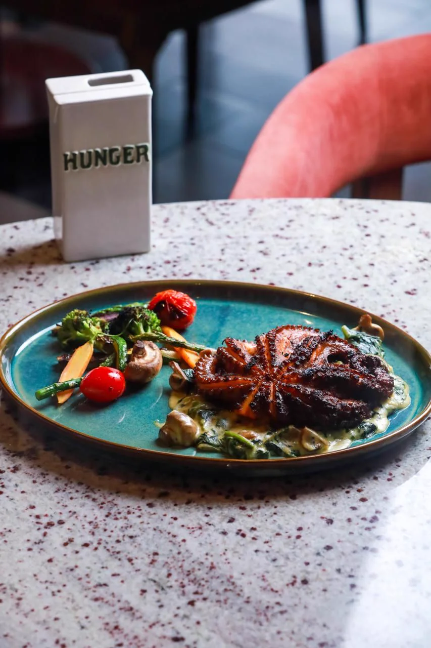 Pulpo adobado al Josper en plato turquesa, con el cartel de la casa detrás.